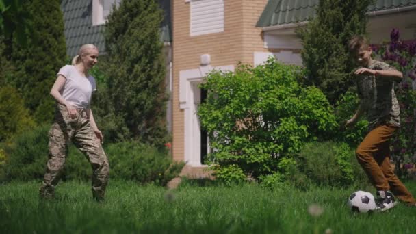 Wide shot vrouwelijke rekruut en tiener jongen voetballen in slow motion op zonnige achtertuin buiten. Happy Caucasian militaire moeder en vrolijke zoon genieten weekend samen op de zomer lente dag. — Stockvideo