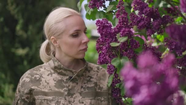 아름다운 금발 여성 병사가 나뭇가지에서 꽃 냄새를 맡으며 카메라를 보고 있습니다. 카키색 위장 제복을 입고 공원에서 느린 동작으로 포즈를 취하는 자신있는 백인 여성의 모습. — 비디오