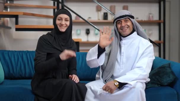 Glada Mellanöstern man och hustru i traditionella kläder vinka leende tittar på kameran talar. Positivt självsäker par sitter på bekväm soffa i mysigt vardagsrum hemma poserar, — Stockvideo
