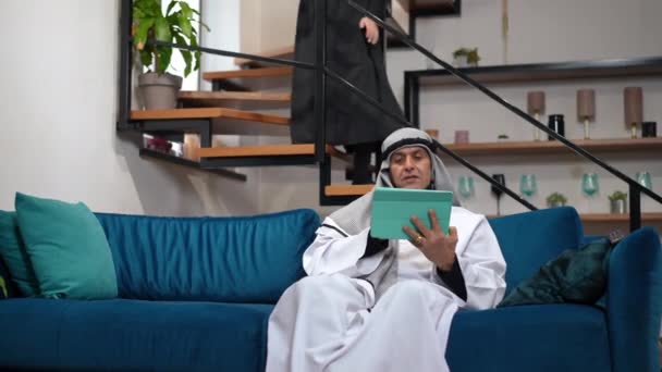 Pria Timur Tengah yang fokus berselancar internet di tablet duduk di ruang tamu yang nyaman sebagai wanita yang tidak dikenal di shayla dan abaya duduk di tangga di latar belakang. Suami dengan istri di dalam rumah. — Stok Video