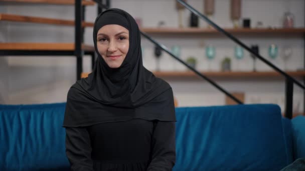 Portret uśmiechniętej pięknej bliskowschodniej kobiety w czarnym hidżabie shayli i abaja siedzącej na kanapie patrzącej w kamerę. Pewna siebie kobieta pozująca w domu po lewej. Koncepcja stylu życia. — Wideo stockowe