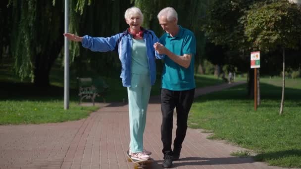 Dolly zastřelil vzrušenou starší žena na skateboardu jako milující muž drží ruce pomáhá manžel. Šťastný kavkazský manžel a manželka cvičení baví v jarním letním parku. Zpomalený pohyb. — Stock video