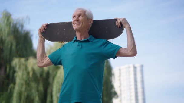 중년의 백인 남성 퇴직자의 행복 한 초상화와 스케이트보드를 어깨에 메고 여름 공원에 서서 주위를 둘러보는 모습. 밝은 날 야외 훈련을 즐기고 있는 자신 감넘치는 회색 머리의 남자. — 비디오