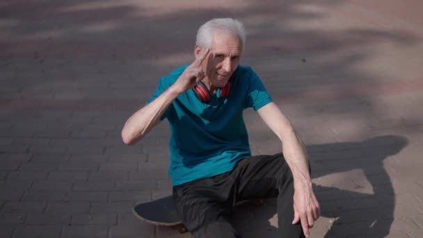 Vue en angle élevé sportif senior positif assis sur skateboard sur une allée ensoleillée du parc saluant au ralenti en regardant la caméra. Joyeux beau caucasien retraité posant en plein air souriant. — Video