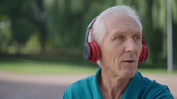 Närbild porträtt av bekymmerslös vit senior man sjunga dans i slow motion lyssna på musik i hörlurar. Avslappnad manlig pensionär njuter av hobby utomhus i sommarparken våren. Hobby livsstil. — Stockvideo