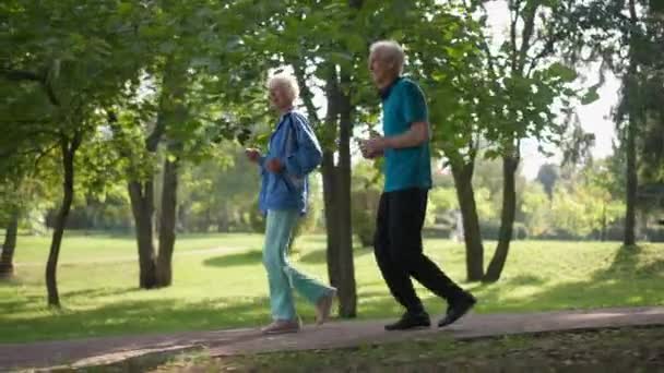 Side view boldog idős pár fut lassított felvételen napsugarak szabadban mosolyogva. Széles lövés magabiztos kaukázusi fitt férfi és nő kocog a napsütésben a tavaszi nyári parkban. Sport és szerelem. — Stock videók