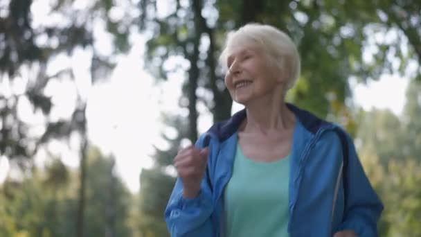 Spännande seniorsportskvinna som joggar i solstrålar i sommarparken och skrattar. Porträtt av glad passform vit pensionär med grått hår i slow motion tittar bort. Individualitet och idrott — Stockvideo