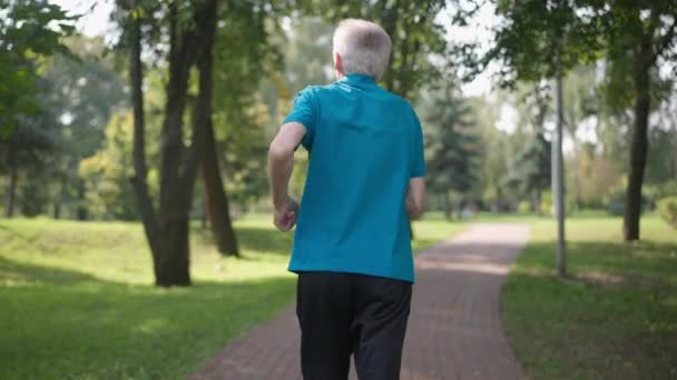 Vue arrière confiant ajustement homme âgé courant dans le parc d'été ensoleillé au ralenti. Suivi de tir Homme caucasien retraité jogging entraînement à l'extérieur le jour du printemps été. Mode de vie sain et longévité. — Video