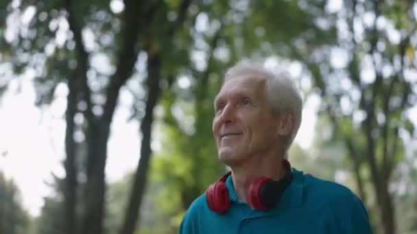 Egy közeli, boldog, idős férfi kocog a szabadban napsugarakban, mosolyogva, félrenéz. Egy magabiztos fehér nyugdíjas portréja, aki lassított felvételen fut a nyári tavaszi parkban. Sport és egészség. — Stock videók