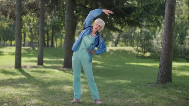 Brede shot fit slanke senior vrouw buigen in slow motion warming-up training in de zomer voorjaar park. Portret van zelfverzekerde blanke gepensioneerde die buiten aan het lachen is. Gezonde levensstijl en vreugde. — Stockvideo