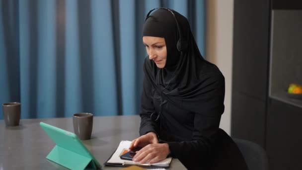 Впевнена розумна жінка з Близького Сходу в традиційному одязі і навушниках, що зустрічається в Інтернеті з серфінгом на планшеті. Розумний вільнодумник розмовляє вдома посміхаючись у мозковий штурм — стокове відео