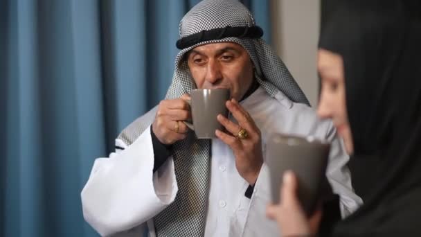 Porträt eines Mannes aus dem Nahen Osten, der den morgendlichen Kaffee genießt und mit seiner Frau lächelt. Liebender glücklicher Mann in traditioneller Kleidung plaudert mit Frau in Küche zu Hause drinnen. — Stockvideo