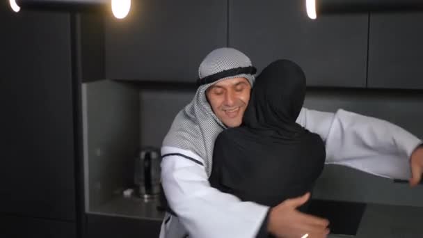 Portret van een gelukkige liefhebbende Midden-Oosterse man in een geruite keffiyeh knuffelende vrouw glimlachend. Knappe man in ghutra omarmen vrouw in shayla staan in de keuken thuis binnen. — Stockvideo