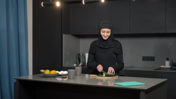 Affascinante sottile donna mediorientale affettare cetriolo per insalata salutare come uomo sorridente che entra in cucina. Positiva coppia felice a casa al chiuso la sera. Stile di vita e concetto di cucina. — Video Stock