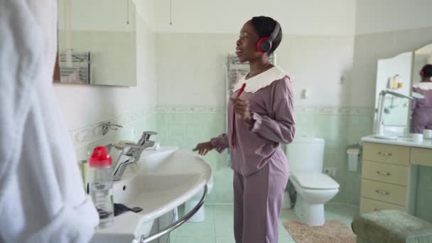Vista lateral alegre jovem mulher magra em pijama dançando em câmera lenta no banheiro admirando a reflexão no espelho. Africano americano milenar senhora em fones de ouvido desfrutando fim de semana dentro de casa. — Vídeo de Stock