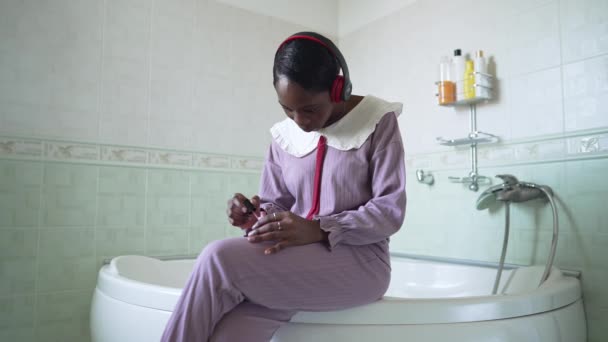 Молода афроамериканська сфокусована жінка, застосовуючи лак для нігтів у повільному русі, сидить на ванній кімнаті у ванній кімнаті. Портрет сконцентрованої стрункої пані в навушниках вранці в приміщенні.. — стокове відео