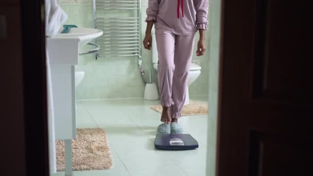 Pedestal shot van slanke jonge Afro-Amerikaanse vrouw stappen op badkamer schalen in slow motion. Slanke millennial dame in pyjama meten gewichtsverlies in de ochtend thuis. — Stockvideo