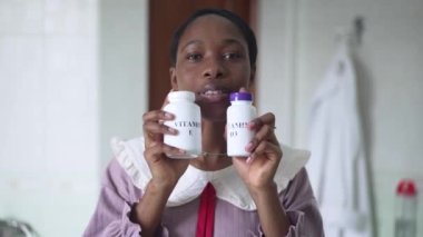Pozitif Afro-Amerikalı kadın, E vitamini ve D3 reklamını banyoda yavaş çekimde yapıyor. Kendine güvenen genç bir kadının portresi. Kameraya bakıyor. İlaç şişelerini gülümserken gösteriyor..