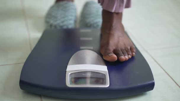 Balanças de banheiro close-up com pés femininos afro-americanos descalços pisando no dispositivo. Jovem magro irreconhecível que mede o peso corporal em casa de manhã. — Vídeo de Stock