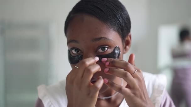 Wajah dekat dari wanita muda Afrika-Amerika terkonsentrasi menerapkan masker mata dalam gerakan lambat menatap kamera. Tampilan depan potret wanita cantik langsing mengurus kulit di pagi hari di kamar mandi — Stok Video