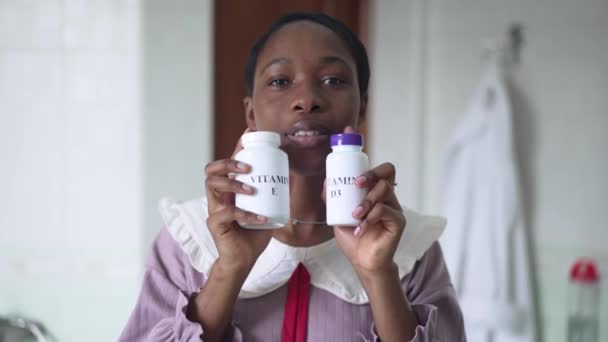 Positieve Afro-Amerikaanse vrouw reclame vitamine E en D3 in slow motion in de badkamer. Portret van zelfverzekerde aantrekkelijke jonge dame op zoek naar camera met pillen flessen glimlachen. — Stockvideo