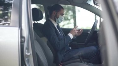 Coronavirus maskeli beyaz adam sürücü koltuğunda oturmuş dezenfektanla ellerini dezenfekte ediyor. Covid-19 salgınında arabadaki zarif, kendine güvenen genç iş adamının portresi..