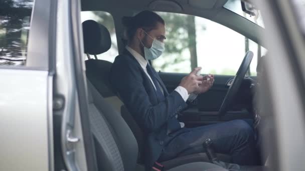 Homem caucasiano em coronavírus máscara facial sentado em motoristas assento desinfetando as mãos com desinfetante. Vista lateral retrato do jovem empresário confiante elegante no carro na pandemia de Covid-19. — Vídeo de Stock