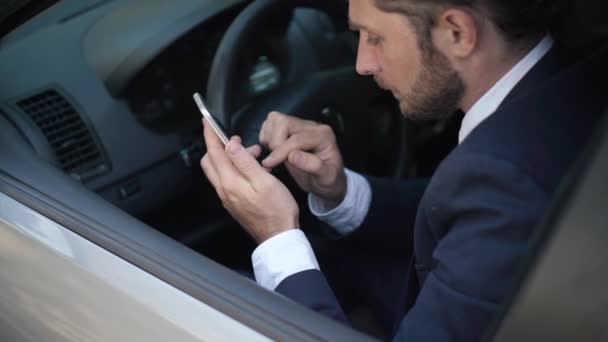 GPSナビゲーションアプリを起動するスマートフォン上のドライバシートテキスト上の高角度ビューの若い男。ハンサムはスマートフォンで車の中で白人ドライバーを集中させた。現代の技術と利便性. — ストック動画
