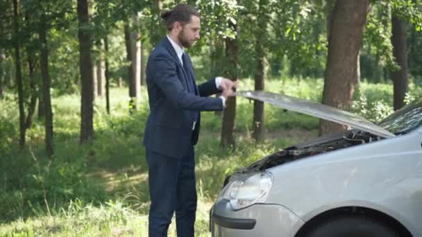 Молодий чоловік, який відкриває автокапот, вдивляючись усередину, думає, що стоїть на сонячному приміському узбіччі дороги. Портрет водія, зосередженого на боці, з розбитим автомобілем на відкритому повітрі. Концепція перегріву двигуна. — стокове відео