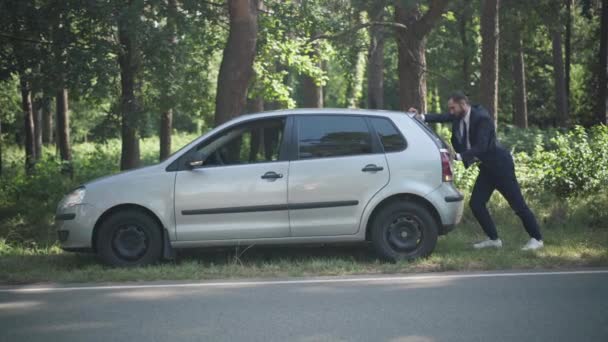 Wide shot man pushing broken car with breakdown lights on on suburban roadside. Young Caucasian guy with vehicle outdoors after accident. Auto industry and lifestyle. — Stock Video