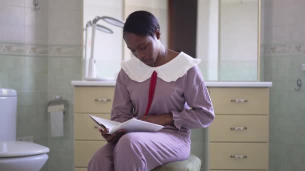 Absorvido sorrindo afro-americano mulher ler livro sentado no banheiro. Retrato de senhora encantadora inteligente em pijama desfrutando de hobby na manhã de fim de semana em casa. Movimento lento. — Vídeo de Stock