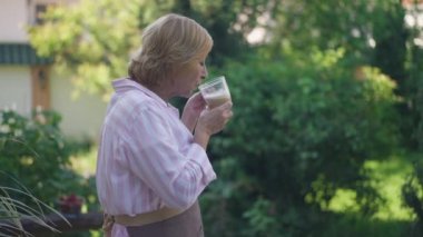 Kendine güvenen yaşlı bir kadının sabah kahvesini yavaş çekimde içerken arka bahçedeki boş vakitlerin tadını çıkarırken yan görüntüsü. Mutlu Kafkas emeklileri gülümseyip cappuccinonun tadını çıkarırken doğaya hayranlık duyarlar..