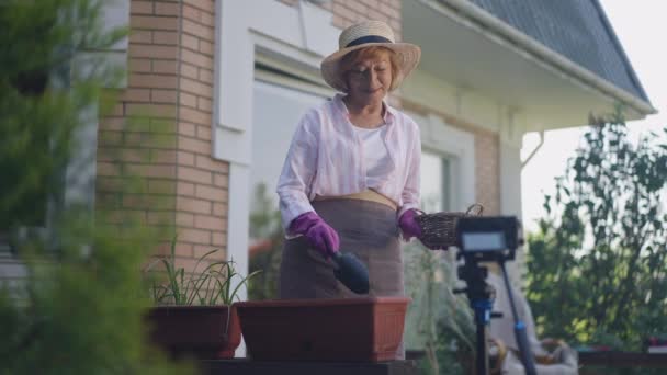 Senior säker kaukasisk kvinna plantera blommor i potten inspelning sociala medier video blogg på bakgård veranda. Porträtt av positiv bloggare pensionär njuter hobby utomhus i slow motion. — Stockvideo