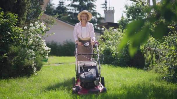 Positiv selbstbewusste Seniorin, die im Sonnenschein mit dem Rasenmäher in Zeitlupe Gras auf dem Hinterhof trimmt. Lächelnde charmante kaukasische Rentnerin genießt sonnigen Sommertag und kümmert sich um den Garten. — Stockvideo