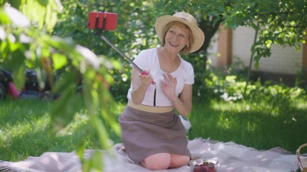 スマートフォンのビデオチャットで有機イチゴを誇る話を振ってワイドショット喜びシニア女性ブロガー。幸せな笑顔の肖像｜白人女性はスローモーションでソーシャルメディアで収穫を自慢. — ストック動画