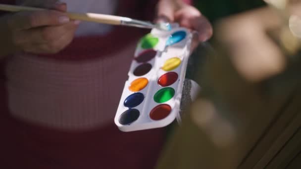 Paleta de pintura de primer plano a la luz del sol con las manos femeninas mayores arrugadas pintando en cámara lenta. Irreconocible talentoso jubilado caucásico disfrutando de hobby en la luz del sol al aire libre. Talento y envejecimiento. — Vídeo de stock