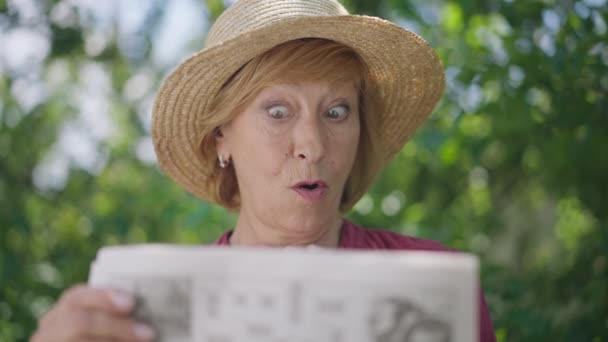 Front view thoughtful embarrassed Caucasian senior woman solving crossword puzzle in slow motion outdoors in sunshine. Portrait of confused retiree with newspaper thinking grimacing. — Stock Video