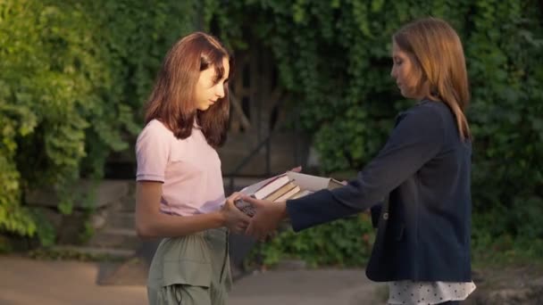 Dvě chytré dospívající dívky si vyměňují knihy ve zpomaleném filmu při západu slunce v jarním letním parku. Boční pohled portrét sebevědomých inteligentních bělošských spolužáků, kteří se scházejí venku ve zlatém slunci. — Stock video
