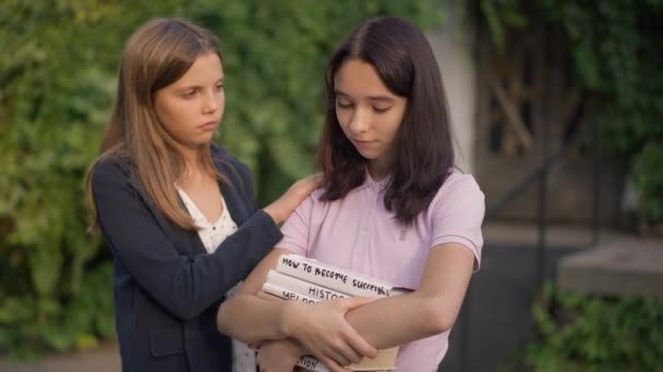 Smutný nerd dospívající dívka s knihami stojí v parku jako přítel laskání rameno uklidňující rozrušený teenager. Empatetický kavkazský dospívající podporující podporující přítele při západu slunce venku. — Stock video