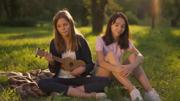 Ευρεία πυροβόλησε ταλαντούχα ευτυχισμένη έφηβη κοπέλα τραγουδώντας ukulele στις ακτίνες του ήλιου ως φίλος ακούγοντας χαμογελώντας. Δύο Καυκάσια κορίτσια απολαμβάνουν το ηλιοβασίλεμα αναψυχής κάθεται στο πράσινο λιβάδι σε εξωτερικούς χώρους πάρκο. — Αρχείο Βίντεο