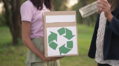 Geri dönüşüm işaretli yakın çekim kutusu ve arka planda yavaş çekimde çöp toplayan bulanık ergen kızlar. Beyaz tenli gönüllü gençler yaz parkını temizliyor..