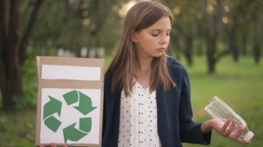 Geri dönüşüm levhası ve kameraya gülümseyen plastik şişeyle kutuya bakan sevimli genç kız. Yazın yaz parkında yavaş çekimde poz veren beyaz bir genç çevrecinin portresi..