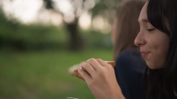 Νεαρή κοπέλα στα δεξιά τρώει σάντουιτς με θολή φίλη στο παρασκήνιο. Θετική χαλαρή Καυκάσιοι έφηβοι απολαμβάνουν πικνίκ στο όμορφο τοπίο. Πρότυπο Mockup. Αργή κίνηση. — Αρχείο Βίντεο