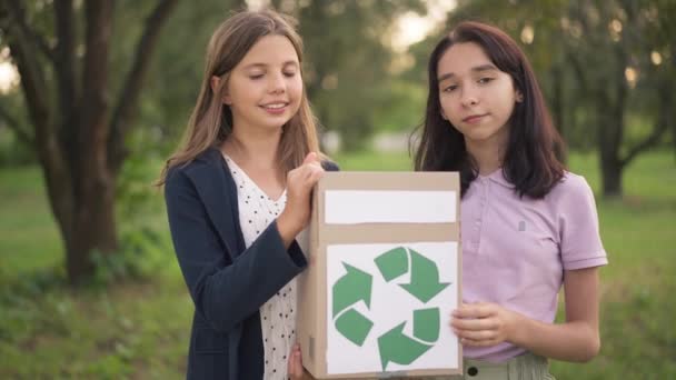 Dos chicas adolescentes caucásicas confiadas posando con caja con cartel de reciclaje verde en el parque de primavera de verano al aire libre. Adolescentes conscientes limpiando la naturaleza al aire libre al atardecer mirando a la cámara sonriendo. — Vídeos de Stock