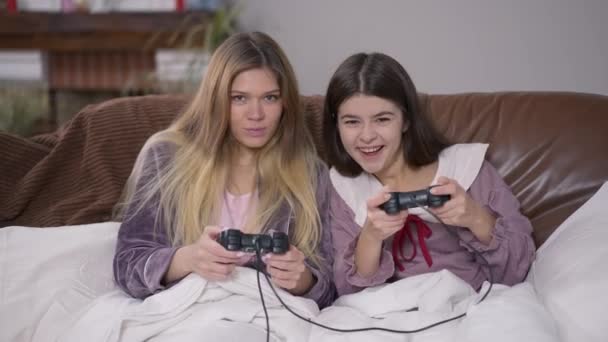 Μπροστά δείτε δύο θετικές νεαρές γυναίκες που παίζουν βιντεοπαιχνίδι με joysticks χαμογελώντας κοιτάζοντας την κάμερα. Καλή ανέμελη Καυκάσιοι φίλοι με πιτζάμες απολαμβάνοντας ελεύθερο Σαββατοκύριακο σε εσωτερικούς χώρους διασκεδάζοντας online gaming. — Αρχείο Βίντεο