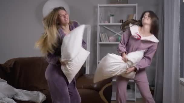 Две веселые кавказские девушки танцуют с подушками на пижамной вечеринке в помещении. Расслабленные уверенные в себе подруги, веселящиеся выходного дня. Счастье и образ жизни. — стоковое видео