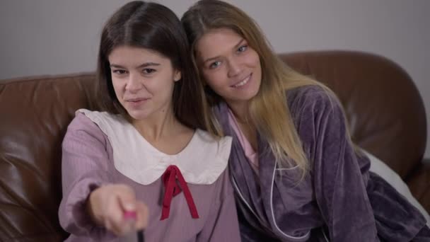 Selfie camera POV of two relaxed young women photographing at pajama party. Cheerful beautiful Caucasian friends posing smiling enjoying leisure indoors sitting on couch. — Stock Video