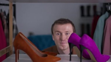 Moda dükkanının rafında, neşeli eşcinsel bir adamın yavaş çekimde ortaya çıkıp saklandığı yakın plan ayakkabılar. Kafkasyalı LGBT elemanı kameraya bakıp gülümseyerek eğleniyor..