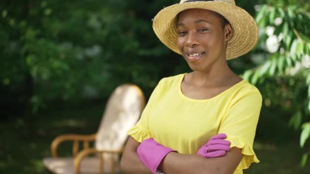 Pewna siebie młoda Afroamerykanka w rękawiczkach ogrodniczych i słomkowym kapeluszu, patrząca na uśmiechniętą kamerę. Portret szczupłego, uroczego ogrodnika pozującego w słoneczny wiosenny letni dzień ze skrzyżowanymi rękami. — Wideo stockowe