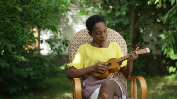 Портрет талановитої афроамериканської жінки, яка грає на укулеле в повільному русі, сидячи на кріслі на задньому дворі. Впевнена, худа молода леді насолоджується хобі на літніх вихідних весни. — стокове відео